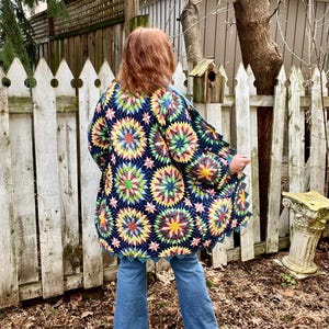 May include: A navy blue, long-sleeved jacket with a colorful starburst pattern. The jacket has a scalloped hem and is worn with blue jeans. The design features various colors, including yellow, pink, green, and blue.