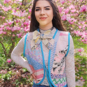 以下が含まれることがあります： ピンクの花が咲いた木の前に立っている女性。彼女は白いレースのシャツに、パッチワークのベストを着ています。ベストは青、ピンク、白の様々な生地で作られており、大きなリボンで結ばれています。