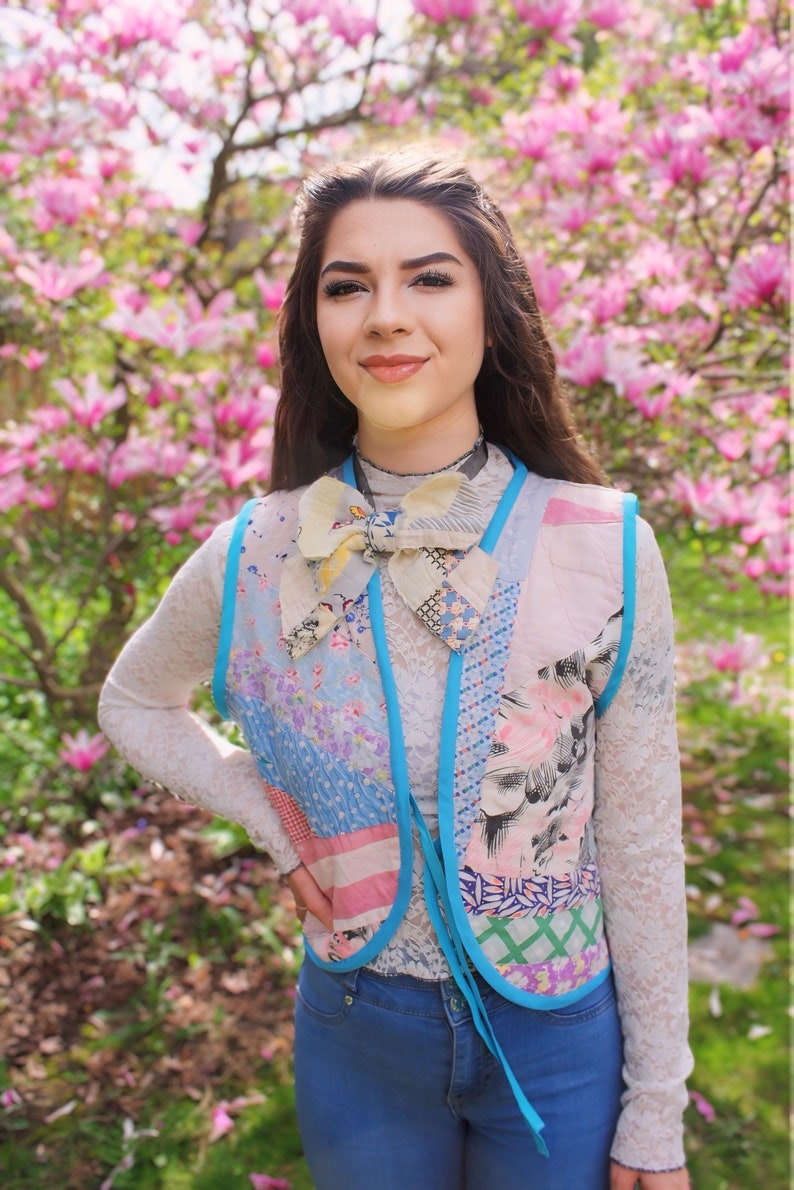 以下が含まれることがあります： ピンクの花が咲いた木の前に立っている女性。彼女は白いレースのシャツに、パッチワークのベストを着ています。ベストは青、ピンク、白の様々な生地で作られており、大きなリボンで結ばれています。