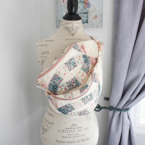 May include: A quilted crossbody bag featuring a patchwork design with floral patterns in shades of pink, green, and cream. The bag has a curved shape, a top zipper closure with metallic hardware, and is displayed on a dress form mannequin.