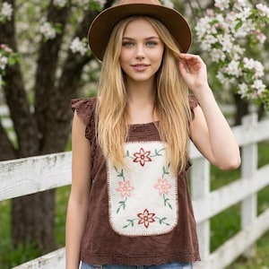 May include: A brown fedora hat and a brown sleeveless top featuring a cream-coloured rectangular panel with embroidered pink and red flowers, green stems, and lace trim on the shoulders and hem, paired with light blue distressed denim jeans.