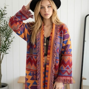May include: A long-sleeved open-front cardigan featuring a vibrant geometric pattern in shades of orange, red, purple, and blue. The garment includes a textured fringe trim along the front opening and sleeve cuffs, creating a distinct layered look.