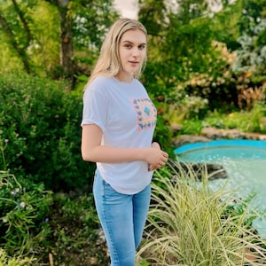 May include: A woman wearing a white t-shirt with a colourful floral design and blue jeans. She is standing in front of a small pond with green plants.