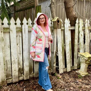 Puede incluir: Chaqueta acolchada con capucha, con un diseño de patchwork en tonos rosa, blanco y verde. La chaqueta tiene forro y ribete rosa. La persona lleva vaqueros azules con rodillas rasgadas. Prenda única.