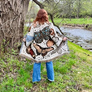 May include: A cream-coloured shawl adorned with butterfly motifs and a fringed border. The shawl showcases diverse butterfly designs in brown, blue, and cream hues. The person is wearing blue jeans near a river.