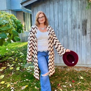 May include: A long, open-front crochet cardigan in cream, brown, and beige chevron pattern. The cardigan is worn over a white lace top and blue jeans. A burgundy hat is held in the hand. The setting is outdoors with a wooden building in the background.