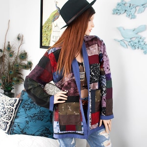 May include: A patchwork jacket in various colours, including red, purple, and black, is worn over a top and blue jeans. The jacket has a blue trim and two pockets. A black hat is worn on the head.