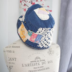 May include: A denim bucket hat with patchwork details and a crocheted lace brim. The hat features various fabric patches in shades of yellow, pink, and blue. The hat is displayed on a mannequin.