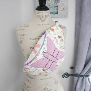 May include: A crossbody bag featuring a cream-coloured fabric with a floral pattern and a large, embroidered pink butterfly design. The bag includes a zip closure with a vibrant, multi-coloured gradient zip tape and a matching pull tab.