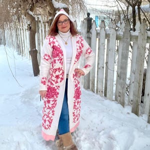 May include: A long, white hooded coat with a pink floral pattern. The coat features pockets and a pink trim. The person is wearing blue jeans and fur-lined boots. The background is a snowy outdoor scene with a white picket fence.