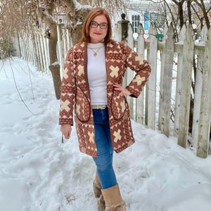 May include: A long, brown and cream-coloured quilted coat with a geometric pattern. The coat has large pockets and is worn over a white top and blue jeans. The person is wearing fur-lined boots and standing in the snow.