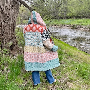 Puede incluir: Un abrigo con capucha de patchwork con una variedad de patrones y colores, incluyendo rojo, turquesa y dorado. El abrigo se usa al aire libre cerca de un río, con vaqueros azules visibles.