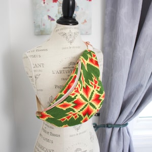 May include: A crescent-shaped crossbody bag featuring a vibrant green, yellow, and red geometric star pattern. The bag has a light-coloured zip closure along the top edge and an adjustable strap, displayed on a mannequin torso.