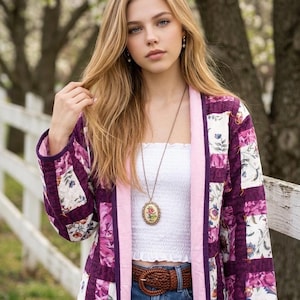 May include: A quilted jacket featuring a patchwork pattern of purple, pink, and white floral fabric, worn over a white smocked tube top and blue denim jeans with a brown braided belt, accessorised with a long oval pendant necklace.