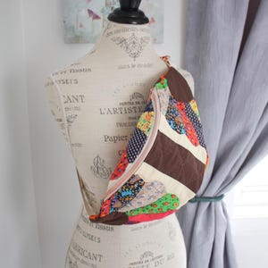 May include: A crescent-shaped crossbody bag featuring a quilted design with a mix of colourful floral, polka dot, and solid brown fabric patches. The bag includes a beige zipper closure and an adjustable strap, displayed on a dress form mannequin.