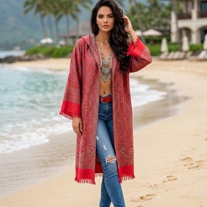 May include: A long-sleeved, hooded red duster coat featuring an intricate paisley pattern in darker red tones, finished with a solid red border and fringe at the hem and cuffs. The garment is worn over blue distressed denim jeans and a red belt.