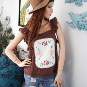 May include: A brown sleeveless top with a white embroidered floral design. The top has a crocheted trim and a square neckline. The model is wearing the top with distressed blue jeans and a brown fedora.