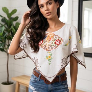 May include: A white cropped poncho featuring a central embroidered graphic of a figure surrounded by orange and red floral motifs, with brown decorative stitching along the edges, paired with blue distressed denim jeans and a brown woven belt.