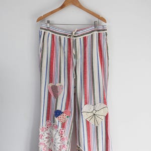 May include: Wide-leg pants featuring a vertical striped pattern in red, blue, green, and white. The pants include a drawstring waist and are decorated with various fabric heart patches, a circular white lace doily, and lace trim at the hems.