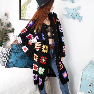 Puede incluir: Cárdigan largo de crochet negro con cuadrados florales coloridos. El abrigo abierto presenta un estilo bohemio. La modelo lleva vaqueros azules y botas de vaquero marrones. Se lleva un sombrero negro en la cabeza.