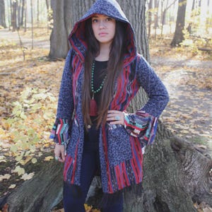 May include: A hooded coat with a unique design. The coat features a blue leopard print hood and sleeves, with red and blue striped panels. The coat has a fringe at the bottom and colorful accents on the sleeves. The person is standing in a forest.
