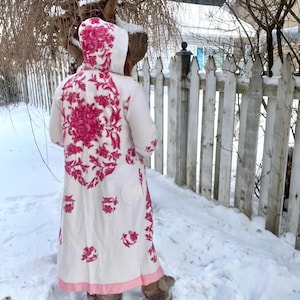 以下が含まれることがあります： フード付きの白いロングコート。背中には大きなピンクの花柄、その他にも小さな花のアクセントがあしらわれています。コートの裾と袖口にはピンクのトリムがあります。白い柵の近くの雪の中に立っている人。