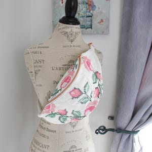 May include: A white fanny pack featuring a cross-stitch pattern of pink flowers and green leaves with a copper-colored zipper. The bag is worn across the torso of a beige dress form printed with French text and decorative flourishes.