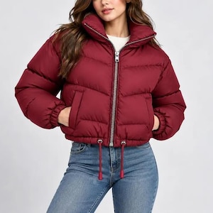 May include: A cropped burgundy puffer jacket featuring a high collar, a silver-toned front zipper, and long sleeves with elasticized cuffs. The jacket includes two side pockets and a drawstring hem with matching burgundy toggles.