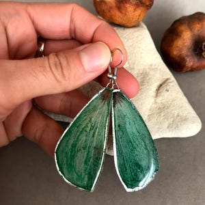 Puede incluir: Un par de pendientes colgantes verdes y plateados con forma de lágrima. Los pendientes están hechos de resina y tienen un efecto marmoleado.