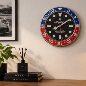 Peut inclure: Une horloge murale ronde conçue pour ressembler au cadran d'une montre Rolex GMT-Master II. Elle présente un cadran noir avec des index blancs, une lunette rouge et bleue avec des chiffres de 24 heures et des aiguilles argentées. Le texte est visible.