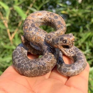 May include: A coiled snake figurine crafted from brown stone with a spotted pattern. The sculpture features a detailed head with an open mouth, visible teeth, and dark circular markings across its entire body surface.