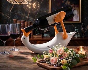 May include: A white ceramic wine bottle holder shaped like a goose with an orange beak and orange feet. The holder supports a dark glass wine bottle horizontally. The goose figure is positioned on a wooden surface next to a floral arrangement.