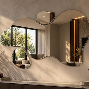 May include: A large, wall-mounted mirror with an irregular, organic, fluid shape and a thin metallic frame. The mirror consists of one main reflective surface and a smaller, separate reflective section positioned above the main piece.