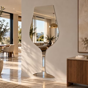 May include: A tall, floor-standing mirror with a unique, asymmetrical silhouette that resembles the shape of a wine glass. The mirror features a reflective glass surface with a smooth, metallic-finished frame, mounted against a plain wall.