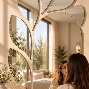 May include: A wall mirror featuring a large central pane with several smaller, leaf-shaped mirror segments attached to the top and left side. The mirror has a smooth, frameless edge and reflects a room interior with a vanity table and accessories.