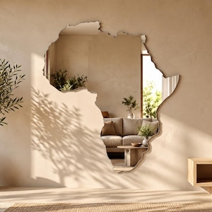 May include: A wall-mounted mirror shaped like the continent of Africa with a smooth, reflective surface. The mirror is positioned on a plain, light-colored wall, reflecting the interior of a room with a sofa and a wooden coffee table.
