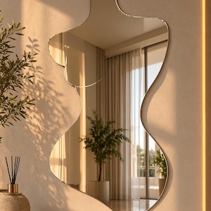 May include: A wall-mounted mirror featuring an irregular, organic silhouette with smooth, flowing curves. The reflective surface captures a room interior with a window, curtains, and a potted plant, creating a distorted, artistic visual effect.