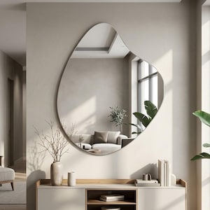 May include: A large, frameless wall mirror with an irregular, organic, pebble-like shape. The reflective surface displays a room interior, including a sofa, a console table, and a potted plant, creating a clear, undistorted reflection.