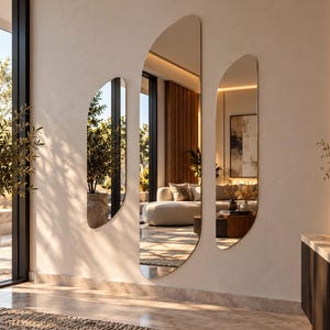 May include: A set of three frameless wall mirrors with irregular, organic, elongated shapes. The mirrors are mounted vertically on a light-colored wall, reflecting a living room interior with a sofa, a coffee table, and natural light.