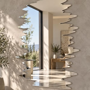 May include: A wall mirror with an irregular, jagged, and organic silhouette. The reflective surface features a smooth, clear finish, while the outer edges are cut into sharp, uneven points that mimic the appearance of a natural fracture.