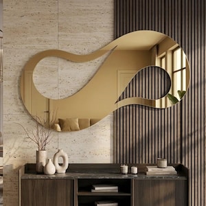 May include: A large, gold-tinted wall mirror with an irregular, fluid, and organic shape. The mirror features a continuous, looping design that creates a hollow center, reflecting the surrounding room and light with a warm, metallic finish.