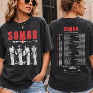 May include: A black short-sleeve t-shirt featuring a red graphic of the band SOMBR on the front with four portraits of band members. The back displays a red tour schedule list with dates, cities, venues, and the text INTERPOL.