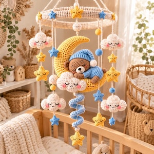 Può includere: Una giostrina per neonati all'uncinetto con una luna crescente gialla centrale e un orsetto marrone che dorme con berretto e pigiama blu. La giostrina include nuvole bianche con guance rosa, stelle gialle e blu appese a una struttura circolare.
