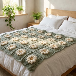 May include: A handmade crochet blanket featuring a repeating pattern of sage green square motifs, each centered with a three-dimensional white flower with a yellow center. The blanket is draped over a white bedspread on a wooden bed frame.