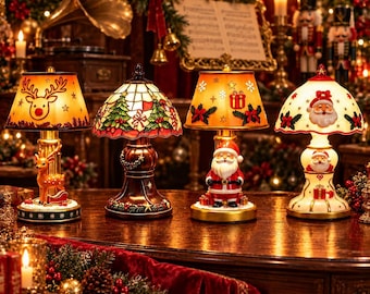 May include: Four decorative Christmas table lamps featuring festive designs. The first lamp has a reindeer base and shade. The second is a stained-glass style lamp with trees. The third features a Santa Claus base. The fourth has a Santa face shade.