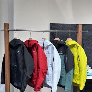 May include: Five Arc'teryx hooded jackets hanging on a metal rack. The jackets are black, red, light blue, a combination of black and dark green, and bright yellow. Each jacket features a front zip, a hood, and the Arc'teryx logo on the chest.