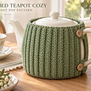 May include: A sage green ribbed crochet teapot cozy featuring two light brown wooden buttons on the side. The text RIBBED TEAPOT COZY CROCHET PDF PATTERN is visible at the top of the image. The cozy is fitted around a white ceramic teapot.