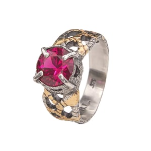 May include: A silver ring with a large, round, magenta gemstone. The band features a textured design with gold accents. The ring is marked with "925" and "B.I."