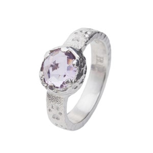 May include: A silver ring featuring a round, faceted, light purple gemstone. The ring has a textured band with small, irregular indentations and a decorative setting around the gemstone. The ring is set against a white background.