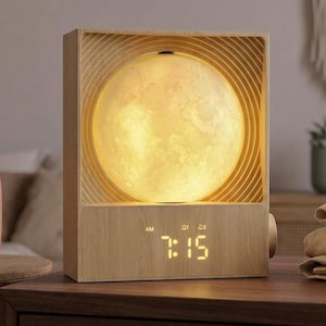 May include: A rectangular light-colored wood alarm clock featuring a circular, glowing moon-like sphere set within a concentric ring frame. The digital display shows the time 7:15 in yellow light, with AM and alarm indicators visible above the numbers.